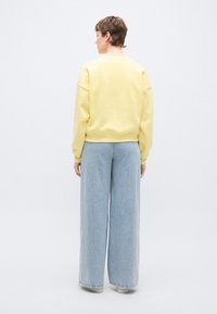 Κίτρινο φούτερ με χαλαρή εφαρμογή και ριγμένα μανίκια, συνδυασμένο με ανοιχτό μπλε τζιν παντελόνι με φαρδιά μπατζάκια. Λεία υφή, απλός σχεδιασμός.