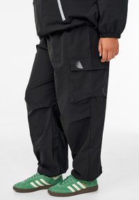 Black cargo pants with a loose fit, featuring a side pocket and a small reflective logo. Paired with green suede sneakers.