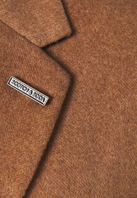 Bruine wollen jas met ingezette kraag en textielstructuur, met een zwart-wit metalen speld met het label "SCOTCH & SODA."