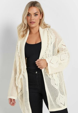 Cardigan a maglia color crema con motivo a traforo, vestibilità oversize, maniche lunghe e dettagli frangiati. Indossato sopra una blusa nera.