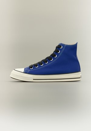 Μπλε ψηλό αθλητικό παπούτσι με μαύρα κορδόνια, λευκή λαστιχένια μύτη και εκρού σόλα με μαύρη ρίγα, εμφανίζεται από το πλάι σε ουδέτερο φόντο.