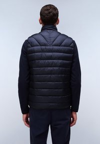Navyblauer gepolsterter Steppweste mit vertikalen Nähten und hohem Kragen, kombiniert mit einem dunklen Pullover, von hinten vor einem schlichten hellen Hintergrund betrachtet.