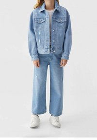 Světle modrá džínová bunda s opranými detaily, zapínáním na knoflíky a dvěma kapsami na prsou, nošená přes bílý top a ladící světle modré kalhoty.
