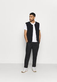 Colete preto com fecho na frente, múltiplos bolsos e um tecido texturizado, usado sobre uma t-shirt branca e combinado com calças escuras e ténis brancos.