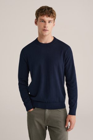 Marineblauer Strickpullover mit langen Ärmeln, gerippten Bündchen und Saum, Rundhalsausschnitt und glatter Textur, kombiniert mit grauen Hosen.