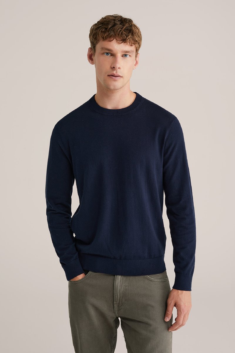 Tamnomodri pleteni pulover z dolgimi rokavi, rebrastimi manšetami in robom, krojnim ovratnikom in gladko teksturo, v kombinaciji s sivimi hlačami.