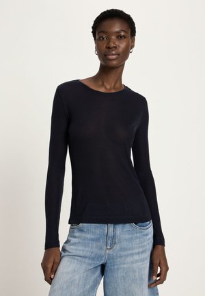 Woman wearing a fitted black long-sleeve top and light blue denim jeans, standing against a plain light background.