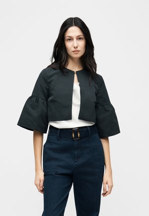 Femme aux longs cheveux noirs portant une veste courte noire à manches évasées, un haut blanc et un pantalon bleu foncé taille haute avec une ceinture.