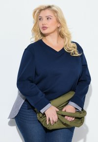 Mjärknäbblå sweatshirt med V-ringning, långa ärmar och randiga mudd. Tillsammans med en olivgrön fluffig väska. Slät textur, avslappnad passform.