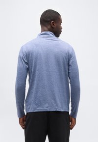 Άνδρας που φαίνεται από πίσω φοράει ανοιχτό μπλε, μακρυμάνικο πουκάμισο με ψηλό γιακά και μαύρο παντελόνι, σε απλό λευκό φόντο.