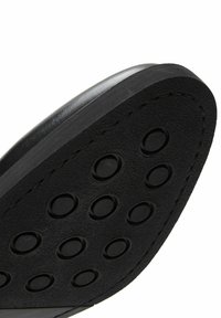 Suela de zapato negra con una superficie texturizada y cortes circulares. El borde superior es liso y hay costuras visibles a lo largo del lado.