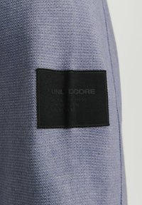 Hellpurpur gestrickter Pullover mit einem schwarzen Gewebelabel und eingeprägtem Text. Die Textur ist weich mit einer leicht groben Webart.