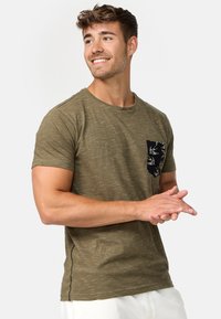Tricou scurt cu mânecă, de culoare verde măsliniu, dintr-o țesătură texturată, cu un buzunar negru ce are un model alb cu păsări. Guler rotund.