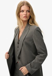 Blazer gris a rayas con un diseño estructurado, solapas de pico y cierre de botón. Presenta un chaleco a juego debajo.