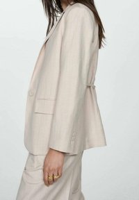 Blazer beige clair avec fines rayures verticales, coupe ajustée, fermeture à un bouton et poches latérales. Tissu doux et texturé, design moderne.