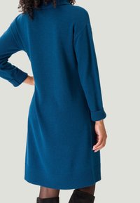 Vestito in maglia teal con colletto alto, polsini a coste e orlo. Realizzato in tessuto morbido, presenta una vestibilità comoda e un design di lunghezza intermedia.