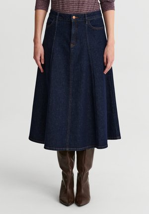 Mujer con una falda vaquera en línea A de color azul oscuro combinada con botas de cuero marrón hasta la rodilla, de pie contra un fondo liso.