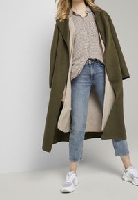 Manteau olive vert avec un large col, doublure intérieure beige, chemise à carreaux, jean bleu et baskets blanches. Tenue décontractée en couches.