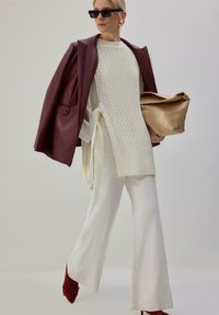 Blazer en cuir bordeaux porté sur un pull blanc en maille torsadée, pantalon blanc évasé et bottines rouges à talons, tenant un sac métallique doré.