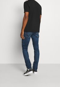 Man som bär en svart t-shirt, blå jeans och svart-vita sneakers, står vänd bortåt mot en ljus enfärgad bakgrund.