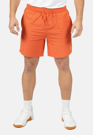 Hombre vistiendo pantalones cortos naranja brillante con cordón, zapatillas deportivas blancas, calcetines blancos y camisa blanca, de pie con los brazos relajados a los lados.