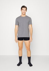 Pilkas trumpas rankovė t-shirt, pritaikytas dizainas. Dėvimi su juodais apatiniais šortais ir tamsiomis kojinėmis, modelis stovi su atsipalaidavusiomis rankomis šonuose, neutralus fonas.