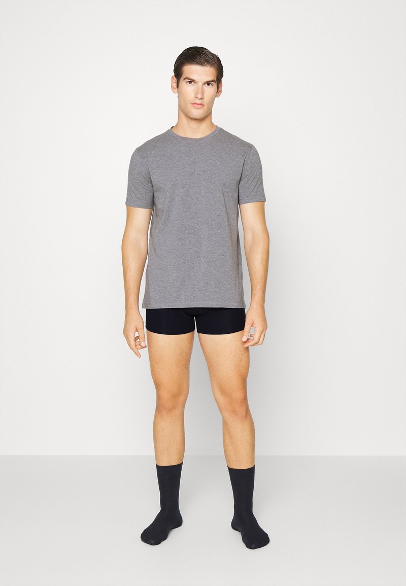 Pilkas trumpas rankovė t-shirt, pritaikytas dizainas. Dėvimi su juodais apatiniais šortais ir tamsiomis kojinėmis, modelis stovi su atsipalaidavusiomis rankomis šonuose, neutralus fonas.