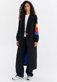 Kobieta o długich kręconych włosach ubrana w czarny długi płaszcz z kolorowym napisem „MORE” na rękawie, białą koszulę, ciemne spodnie i srebrne trampki, stojąca na białym tle.