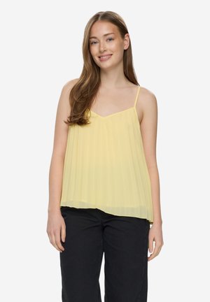 Mladá žena s dlouhými hnědými vlasy, která má na sobě žlutý plisovaný top bez rukávů a černé kalhoty, mírně se usmívá a stojí před jednobarevným pozadím.