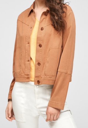 Femme portant une veste beige boutonnée sur une chemise jaune et un pantalon blanc avec une poche zippée, visible du cou jusqu'aux hanches.