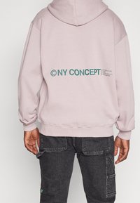 Sudadera con capucha de color rosa claro y ajuste holgado, con gráficos de texto en verde en la espalda. Puños y cinturilla acanalados con un acabado texturizado.