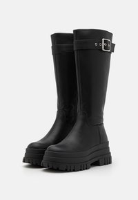 Botas de cuero negro hasta la rodilla con una suela plana gruesa, que presentan una gran hebilla plateada y un patrón de relieve en la suela.
