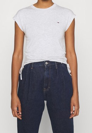 Person trägt ein hellgraues, ärmelloses Oberteil mit seitlichen Bindebändern und dunkle, hoch taillierte Jeans aus Denim, steht vor einem einfarbigen Hintergrund.