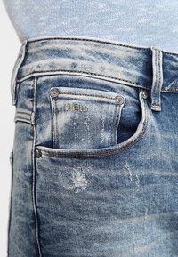 Jeans de mezclilla azul claro con una textura deslavada, que presentan un pequeño bolsillo frontal y costuras visibles. Detalles desgastados cerca de la cinturilla.