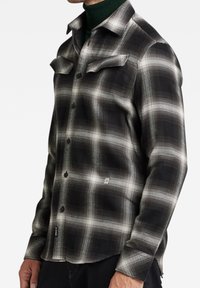 Camicia a quadri bianca e nera con due tasche sul petto, motivo a spina di pesce e orlo curvo. Presenta bottoni neri e un dettaglio del logo.