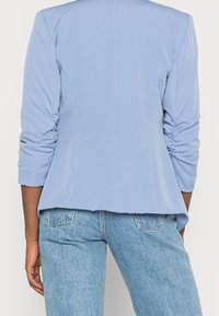 Svetle modrý blejzr s nařasenými rukávy, hladkou texturou, dobře padnoucím střihem a zakřiveným lemem. Spárován se světlomodrými džíny, které mají zadní kapsy.