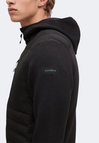 Jeune homme portant une veste matelassée noire avec capuche, montrant le haut du torse et le bras avec un logo Icepeak visible sur la manche.