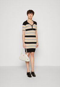 Abito a righe in maglia nero, beige e bianco con maniche corte, scollo a v e dettaglio con bottoni. Indossato con una borsa bianca texturizzata e zoccoli neri.