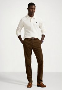 Bela polo majica z dolgimi rokavi in ovratnikom, rjave chino hlače, rjavi usnjeni čevlji in rjav pas. Klasičen kroj z diskretno logotipno podrobnostjo.