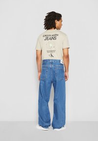 Bēša T-krekls ar melnu grafiku, kombinēts ar brīviem zilajiem džinsiem. Džinsiem ir balts logo uzlīme aizmugurē.