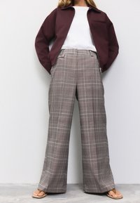 Pantaloni a quadri marroni e grigi con accenti rossi; design a vita alta e gamba larga; abbinati a una giacca borgogna e una maglia bianca.