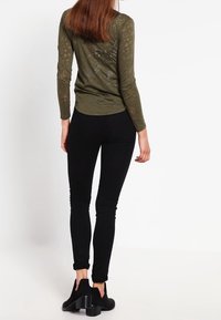 Olivgrön långärmad topp med ett subtilt mönster, tillsammans med svarta slim-fit byxor och svarta ankelskor på en neutral bakgrund.