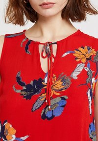 Czerwona, bezrękawnikowa bluzka z kwiecistym haftem w kolorze niebieskim, pomarańczowym i zielonym. Posiada dekolt w kształcie dziurki z wiązaniem. Lekki materiał.