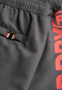 Grijze sportshorts met een elastische tailleband, een mesh zak en een grote rode "SUP" graphic. Het label is oranje, wit en zwart gestreept.