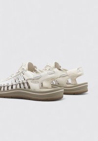 Sandalias de color beige claro con parte superior de tejido trenzado, correa trasera acolchada y suela amortiguada. Presenta el logotipo de la marca en el lateral y una suela texturizada.