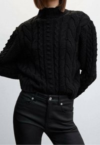 Černý pletený svetr s kabelovým vzorem, vysokým límcem, silnou texturou a volnějším střihem, v kombinaci s černými přiléhavými džíny s knoflíkovým zapínáním a kapsami.