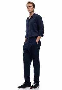 Camicia blu navy con bottoni e maniche arrotolate, abbinata a pantaloni blu navy su misura e sneakers scure con lacci bianchi e suola texturizzata.