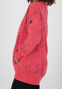 Felpa rossa con motivi floreali neri a contorno, vestibilità oversize, maniche lunghe, polsini a costine e una tasca laterale. Tessuto morbido.