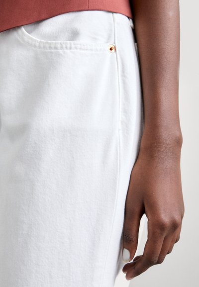 Pantalones de denim blanco con un bolsillo lateral y un detalle de tachuela dorada. La tela tiene una textura sutil, contrastada por una blusa de color óxido.