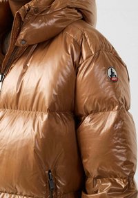 Chaqueta acolchada marrón y brillante con capucha, que cuenta con una cremallera y un parche con logotipo en el hombro izquierdo, caracterizada por una textura acolchada.